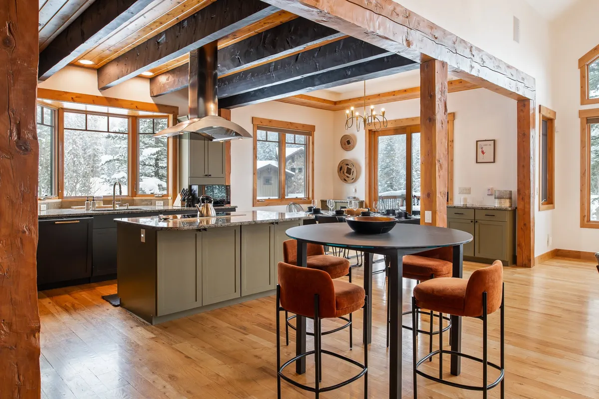 This image presents a warm and cozy kitchen scene featuring a spacious kitchen island with marble or granite countertops, four bar stools, large windows offering views of the snowy landscape, wooden beams, and a striking chandelier. The layout encourages both culinary creativity and social interaction, perfectly aligning with the communal kitchen narrative.