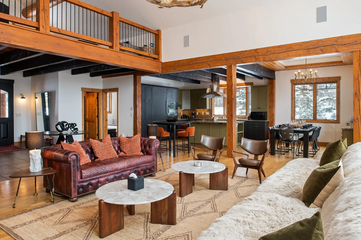 A wide-angle view of the open-concept living and dining area, featuring a leather sofa, exposed wooden beams, a kitchen island, and expansive windows that allow natural light and mountain views to fill the space. The setup encourages conversation, relaxation, and shared experiences.