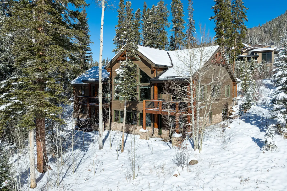 A striking exterior view of the log cabin-style home nestled in a snow-covered landscape, surrounded by tall evergreen trees. The inviting porch hints at relaxed gatherings and cozy moments, while the expanse of snow suggests opportunities for outdoor group activities.