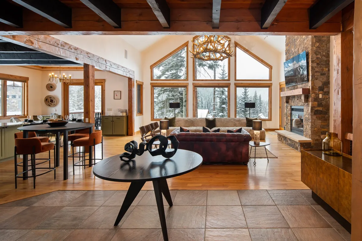 A warm, inviting living area featuring plush seating, a stone fireplace, a dining space, and large windows overlooking a scenic, snow-dusted landscape. The arrangement encourages relaxation and connection, with comfortable furniture and soft lighting creating an atmosphere perfect for group gatherings or cozy evenings.