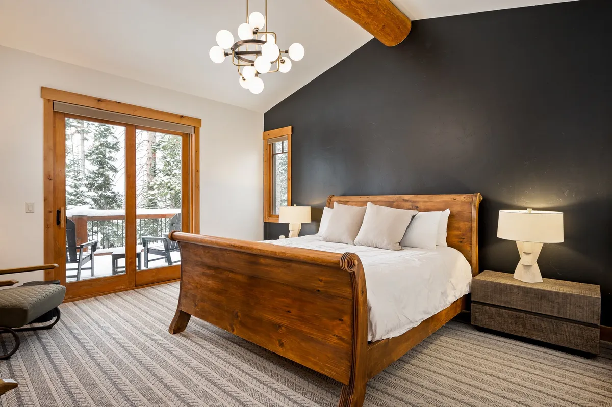 A spacious master bedroom featuring a wooden sleigh bed, large windows revealing a tranquil forest view, a modern chandelier, and a textured area rug. The room exudes a sense of serenity and privacy, ideal for unwinding after a day of adventure.