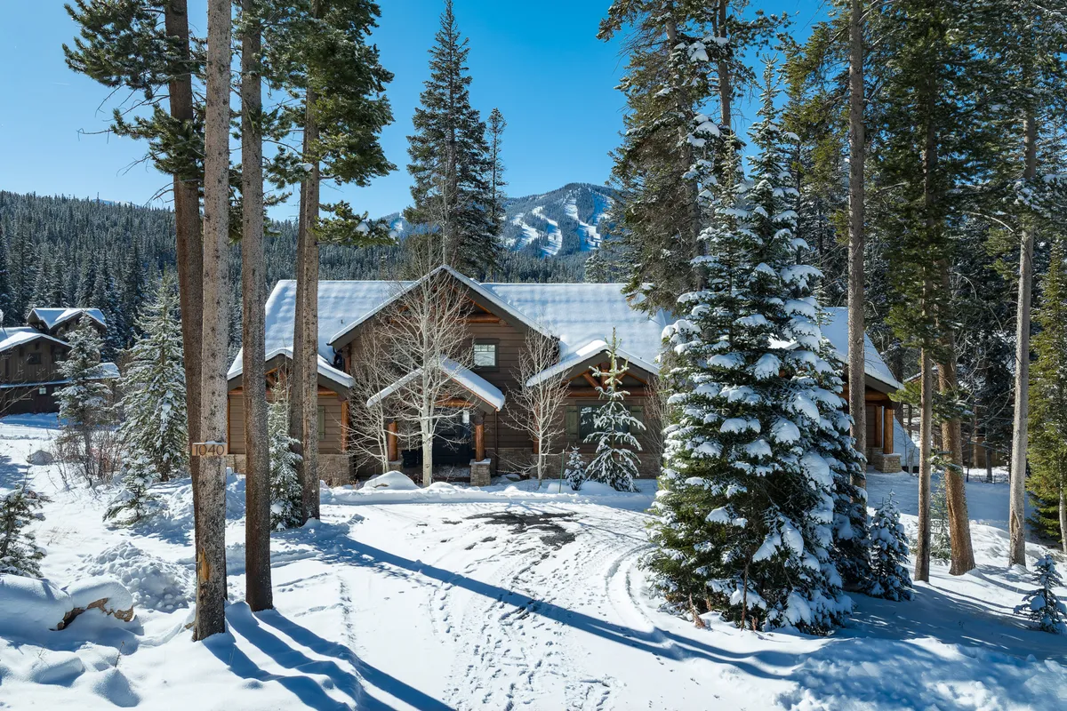 A cozy log cabin nestled among snow-covered trees with a stunning mountain backdrop. The scene evokes a tranquil and inviting winter wonderland, perfect for showcasing winter and early spring activities such as skiing, snowboarding, and enjoying the natural beauty from the porch.