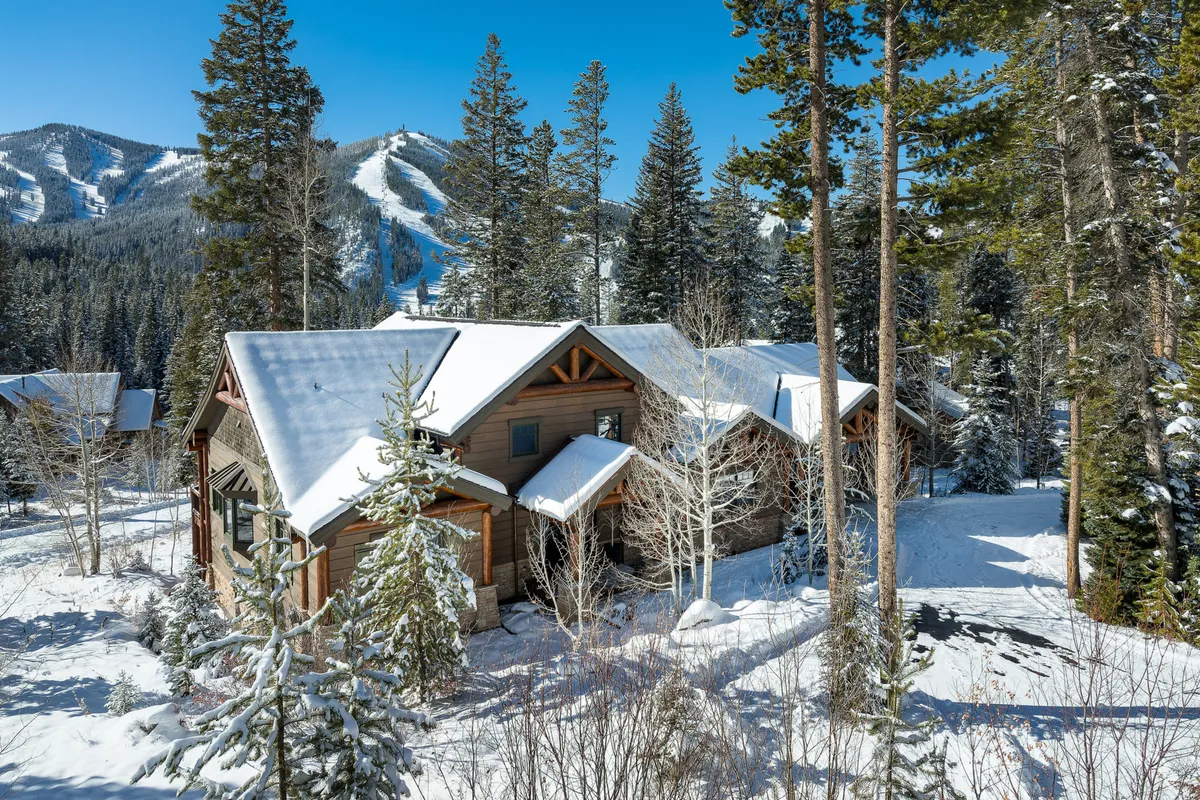 The image showcases a luxurious single-family home blanketed in snow, surrounded by towering pine trees, with majestic mountains rising in the background. The cozy, inviting glow from the windows hints at warmth within, while the natural setting promises adventure right outside the door.