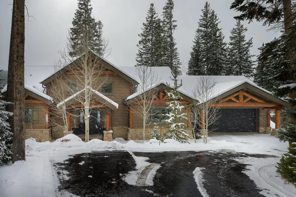 A picturesque log cabin with a snow-covered roof sits among tall evergreen trees. Tire tracks in the driveway hint at recent arrivals, while the serene setting suggests privacy and tranquility. The image emphasizes connection to the outdoors and the inviting warmth of the cabin after winter activities.