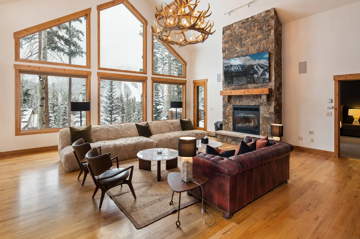 A stunning great room with soaring ceilings and expansive floor-to-ceiling windows that frame panoramic mountain vistas. Centered around a commanding stone fireplace and accented by a unique chandelier, the room is filled with natural light and features plush, comfortable seating ideal for group gatherings.