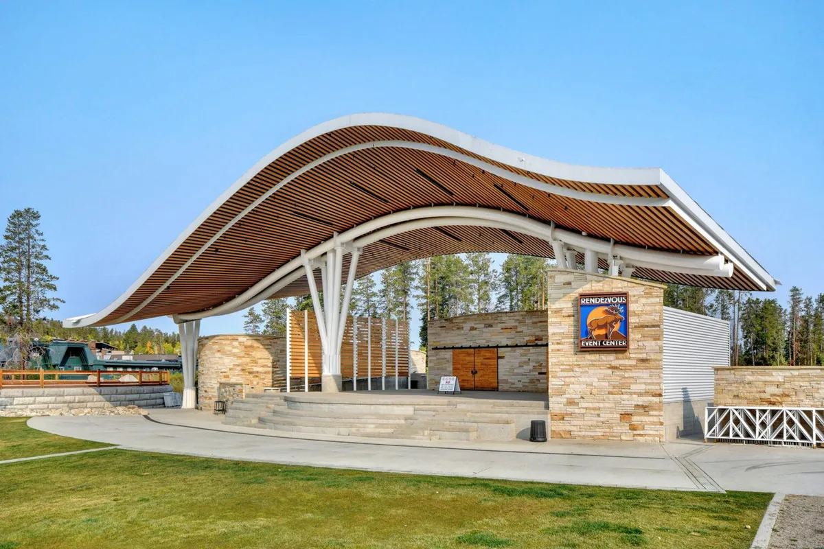 An outdoor venue with a curved roof structure and stone facade, event signage displayed, and space set up for outdoor performances and gatherings. The area is ready for concerts, community events, and picnics, with a clear focus on summer activities and a connection to the local arts scene.