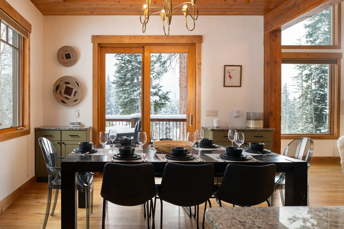 A beautifully set dining room with a large wooden table for eight, surrounded by ample windows showcasing snow-covered trees and featuring a wooden ceiling and a prominent chandelier. The mood is inviting, perfect for family-style feasts and laughter-filled gatherings as described in the section.
