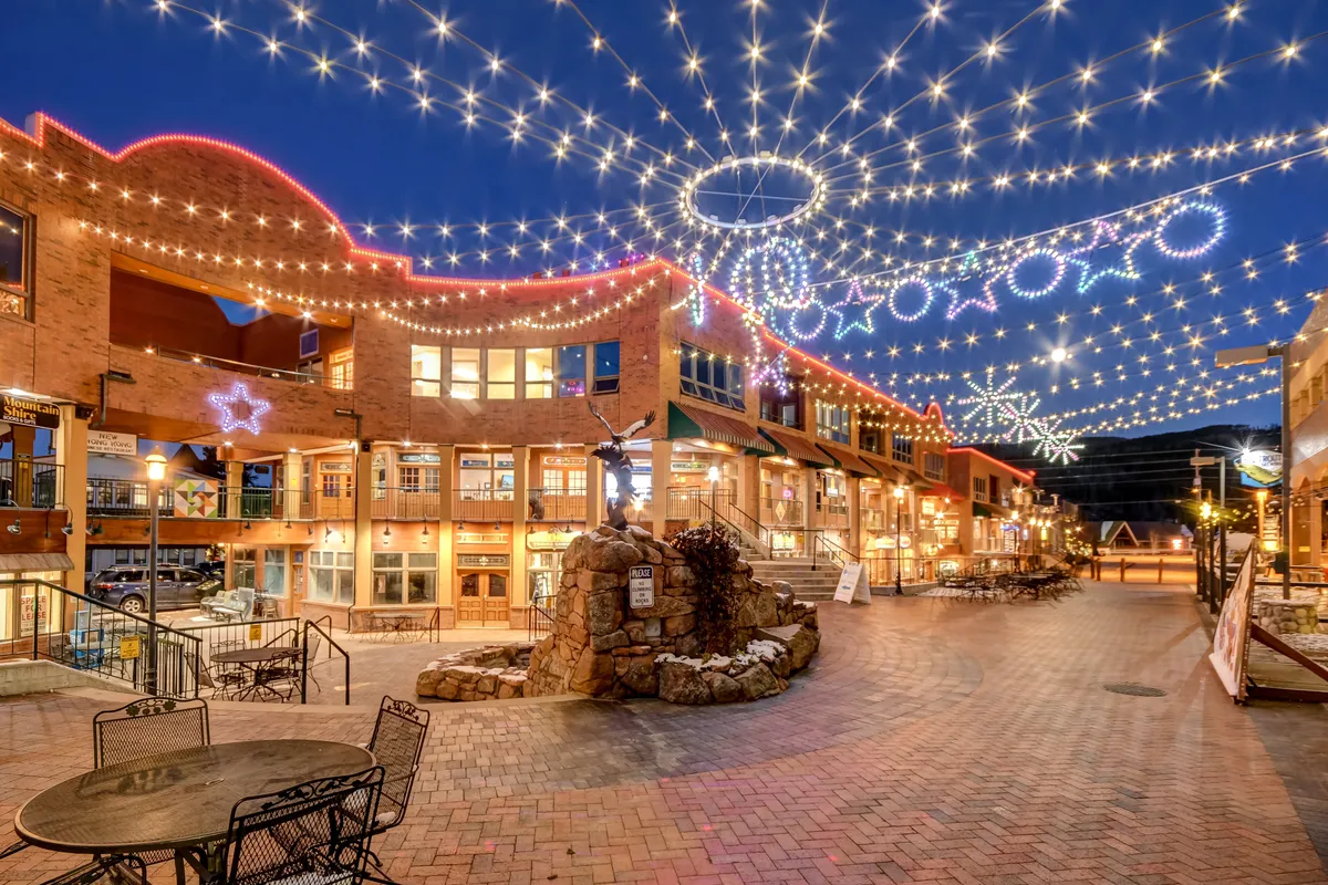 A lively outdoor plaza adorned with string lights and decorative stars above, with a stone fountain and people dining, shopping, and socializing. The scene is festive and bustling, embodying community events and the vibrant downtown atmosphere present in late spring, summer, and fall.